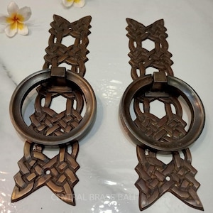 以下が含まれることがあります： リングプルと織り模様が施された、装飾的な真鍮製のドアハンドル2個。ハンドルは星形をしていて、濃い茶色の仕上げになっています。