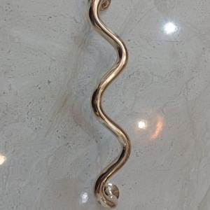 Op de afbeelding: Een goudkleurige, golvende deurklink. De klink heeft een uniek, gebogen ontwerp, met afgeronde uiteinden voor een gemakkelijke grip. De klink is gemaakt van een glanzend, metalen materiaal en is waarschijnlijk voor een kast of deur.