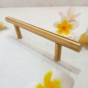 Könnte beinhalten: Ein goldfarbener, zylindrischer Schrankgriff mit zwei vertikalen Pfosten. Die Hardware befindet sich auf einer weißen Oberfläche, mit mehreren weißen und gelben Frangipani-Blüten. Der Text "CENTRAL BRASS BALI" ist sichtbar.
