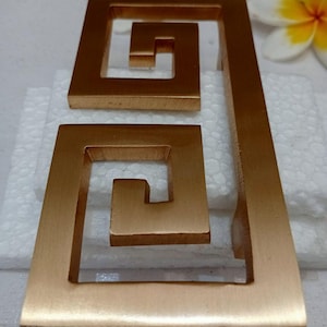 May include: Two gold-colored, geometric drawer pulls with a Greek key design. The pulls are rectangular with a spiral pattern inside. The image also includes two white and yellow flowers. The text "CENTRAL BRASS BALL" is visible at the bottom.