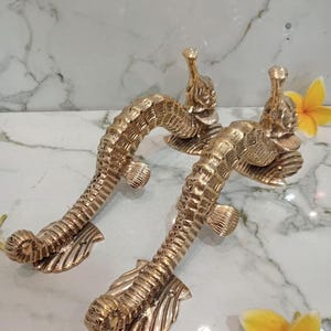Op de afbeelding: Twee messing deurklinken in de vorm van zeepaardjes. De handgrepen zijn gedetailleerd met schubben en vinnen en hebben een gebogen vorm. De handgrepen liggen op een wit marmeren oppervlak, met gele bloemen op de achtergrond. De tekst "CENTRAL BRASS BALI" is zichtbaar.