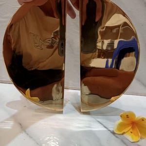 Peut inclure: Deux poignées de porte en laiton poli en forme de demi-lune. Les poignées sont de couleur or brillant et reflètent l'environnement. Le texte "CENTRAL BRASS BALI" est visible en haut à droite. Une fleur jaune à pointes blanches est en bas à droite.