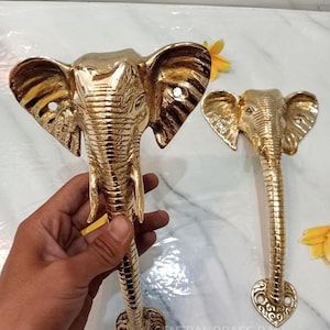 May include: Two polished gold-coloured brass door handles shaped like elephant heads. Each handle features a detailed elephant face with large ears, tusks, and a long trunk that extends downward to a decorative heart-shaped base with screw holes.