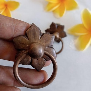 May include: Close-up of two bronze-colored flower-shaped drawer pulls with circular handles. The pulls have a floral design with a central sphere. The background includes yellow flowers.