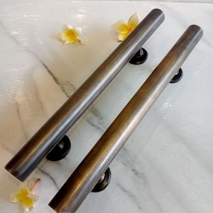 Op de afbeelding: Twee bronzen, cilindrische deurklinken met zwarte montageplaten. De handgrepen zijn diagonaal geplaatst op een wit marmeren oppervlak, met gele en witte bloemen eromheen. Ze hebben een rustieke, verouderde uitstraling.