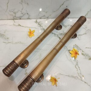 May include: Two bronze-colored door handles with decorative etched ends and circular mounting plates. The handles are cylindrical and taper slightly. The background is a white marble surface with yellow flowers. The text "CENTRAL BRASS BALI" is visible.