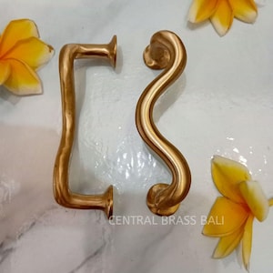 May include: Two gold-toned metal door handles. One handle is a simple rectangular shape with rounded edges. The other handle is a curved, S-shaped design. The handles are on a white surface.