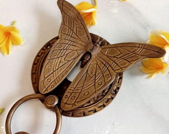 May include: A brass door knocker shaped like a butterfly with intricate details. The knocker has a ring handle and is mounted on a circular base with a decorative border.