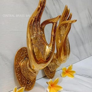 Peut inclure: Deux sculptures murales en laiton doré en forme de mains, avec des détails complexes. Les mains sont dans un geste de bénédiction ou de méditation. Le texte "CENTRAL BRASS BALI" est visible. Des fleurs jaunes sont éparpillées.