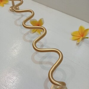 May include: Gold-toned, wavy, metal door handle with a round base. The handle is attached to a white wall.