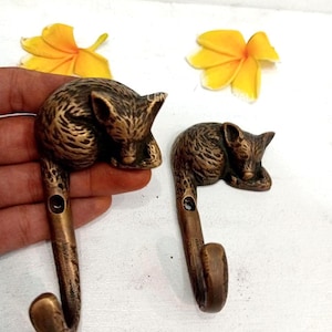 May include: Two bronze-colored metal wall hooks, each featuring a sleeping fox design. The hooks are curved and have a small hole for mounting. The fox figures are detailed with textured fur. Yellow flowers are in the background.