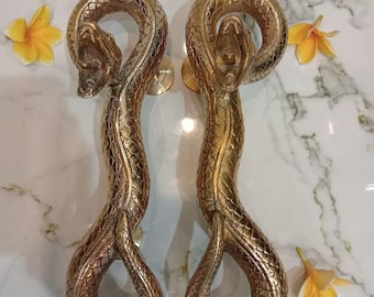 May include: Two brass snake-shaped door handles. The handles are detailed with scales and have open-mouthed snake heads. The handles are gold in color and are displayed on a marble surface with yellow flowers. The text "CENTRAL BRASS BALI" is at the bottom.