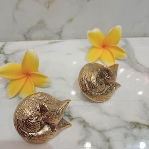 May include: Two gold fox figurines and two yellow and white plumeria flowers are arranged on a marble surface. The fox sculptures are detailed and curled up. The flowers add a touch of colour.