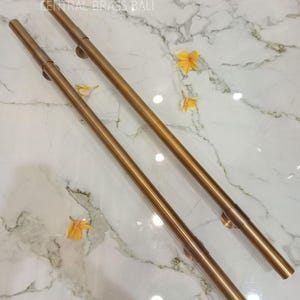 Può includere: Due corrimano in metallo color bronzo con un design cilindrico. I corrimano sono posizionati diagonalmente su una superficie di marmo bianco, con il testo "CENTRAL BRASS BALI" visibile. Fiori gialli sparsi.