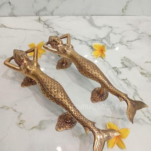 May include: Two brass mermaid door handles. Each handle features a mermaid with her arms behind her head and a scaled tail. The handles are golden in color and are placed on a white marble surface with yellow flowers.