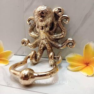 Op de afbeelding: Een bronzen octopusvormige deurklopper. De klopper heeft een gedetailleerd octopusontwerp met gekrulde tentakels en een ringvormige handgreep. De tekst "CENTRAL BRASS BALI" is onderaan zichtbaar. Gele bloemen staan aan weerszijden.