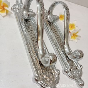 May include: Two silver door handles with ornate designs and a curved top. The handles are mounted on rectangular plates with a textured surface. The plates have a decorative border and a central design.