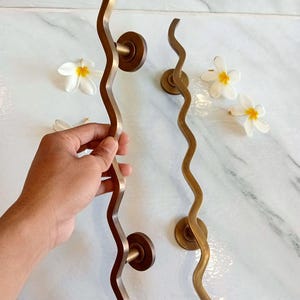 May include: Two wavy brass door handles with round knobs. The handles are a warm gold color and have a smooth, polished finish. The handles are shown against a white marble background.
