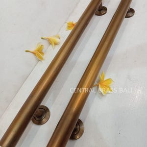 Puede incluir: Dos tiradores de puerta largos de latón pulido con extremos redondos y placas base circulares. Los tiradores están sobre un fondo blanco con flores amarillas.