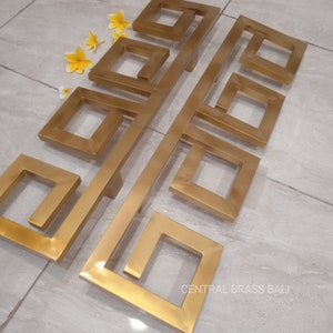 Puede incluir: Conjunto de seis tiradores de puerta de metal dorado con un diseño geométrico cuadrado. Los tiradores están dispuestos en dos filas, con tres tiradores en cada fila. Los tiradores están hechos de metal sólido y tienen un acabado liso y pulido. Los tiradores están etiquetados "CENTRAL BRASS BALI".