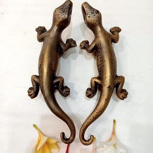 May include: A pair of bronze-toned metal door handles shaped like lizards, featuring textured skin details, curved tails, and splayed feet. The lizard figures are positioned vertically, facing each other, with small floral accents placed below them.