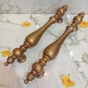 May include: Two elongated brass door handles featuring a smooth, curved central body and decorative spherical accents at both ends. The metal has a warm, brushed gold finish with a polished sheen, showcasing a symmetrical design with rounded finials.