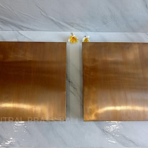 Könnte beinhalten: Zwei quadratische Metallplatten in Kupferfarbe. Die Platten haben eine gebürstete Oberfläche, die das Licht mit einem warmen, metallischen Glanz reflektiert. Die Platten werden auf einer weiß-grauen marmorähnlichen Oberfläche präsentiert. Der Text "CENTRAL BRASS BALI" ist unten sichtbar.