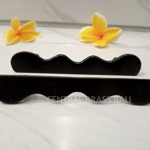 May include: Two black wooden decorative pieces with a wavy design, possibly for a shelf or display. The text "CENTRAL BRASS BALL" is visible. Two yellow flowers are in the background.