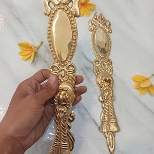 Puede incluir: Dos manijas de puerta de latón dorado ornamentadas. Cada manija presenta un centro ovalado, detalles intrincados y una parte superior e inferior decorativas. Las manijas se exhiben sobre una superficie de mármol con flores amarillas.