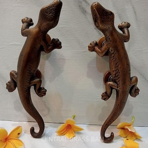 Peut inclure: Deux poignées de porte en laiton antique en forme de gecko avec une finition texturée détaillée. Les poignées sont en forme de geckos avec leurs queues enroulées. Les poignées sont présentées sur un fond de marbre blanc avec des fleurs jaunes.