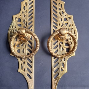 May include: Pair of ornate brass door handles. Each handle features a decorative, openwork design with a textured ring pull. The brass has a warm, golden hue, and the handles are set against a dark background.