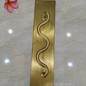 Könnte beinhalten: Ein goldfarbener Türgriff mit wellenförmigem Design. Der Griff ist auf einer rechteckigen Platte mit gebürsteter Oberfläche montiert. Die Platte hat vier kleine Löcher für Montageschrauben. Der Text "CENTRAL BRASS BALI" ist auf der Platte aufgedruckt.