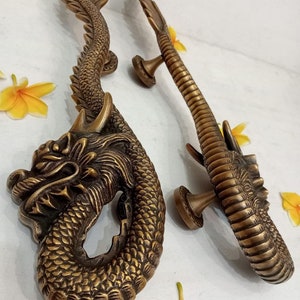 May include: A pair of ornate brass door handles shaped like dragons. The handles are detailed with scales and have a gold finish. The text "CENTRAL BRASS BALI" is visible in the top left corner of the image.