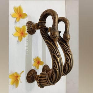 Puede incluir: Un par de tiradores de puerta de latón adornados con forma de cisnes con tallas detalladas. Los tiradores están montados en una pared blanca.