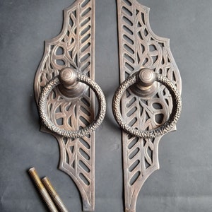 Puede incluir: Dos tiradores de puerta de color bronce con intrincados diseños calados de hojas y vides. Cada tirador tiene un asa de anillo texturizada. Se incluyen dos pernos y tuercas. El herraje es probablemente para un armario o una puerta.
