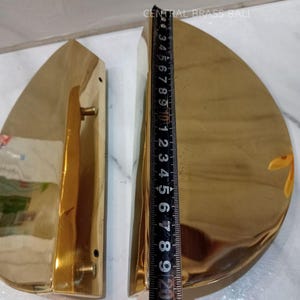 Puede incluir: Dos manijas de puerta de latón pulido, cada una con forma de lágrima y un pequeño mango. Una regla muestra que las manijas miden aproximadamente 25 cm de largo. El texto "CENTRAL BRASS BALI" es visible.