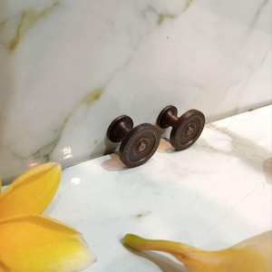 May include: Two antique bronze cabinet knobs with a circular design. The knobs are dark brown and sit on a white and light green marble surface. A yellow flower is in the foreground.