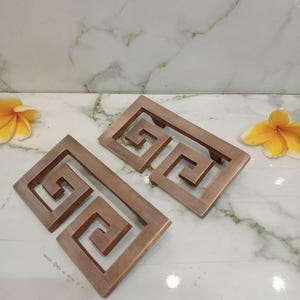 May include: Two rectangular cabinet pulls with a metallic copper finish, each featuring an open-frame design with a repeating geometric Greek key pattern. The hardware pieces are positioned at an angle, showcasing the brushed texture of the metal.