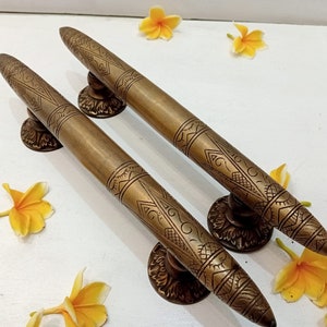 May include: A pair of ornate brass door handles with intricate designs. The handles are long and slender, with a pointed end. They are mounted on a white surface.