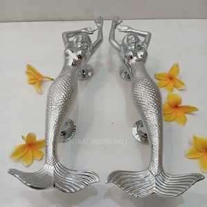 May include: Two silver mermaid door handles with a textured, scaly finish. The mermaids are in a reclining pose with their arms raised above their heads. The handles are mounted on a white background with yellow flowers.