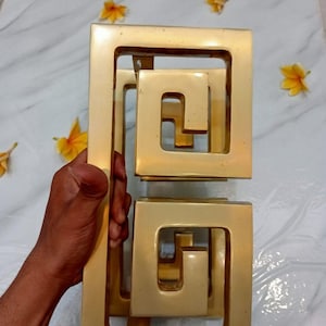 Puede incluir: Pomo de puerta de metal dorado con un diseño geométrico. El pomo está hecho de varios cuadrados entrelazados. El pomo está etiquetado como "CENTRAL BRASS BALI".