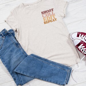 Könnte beinhalten: Ein beiges T-Shirt mit dem Aufdruck "SHOOT CULL EDIT REPEAT" auf der Vorderseite. Das Shirt wird mit einer blauen Jeans und roten Turnschuhen kombiniert.