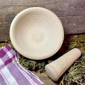 May include: A beige ceramic mortar and pestle set. The mortar is a bowl with a textured interior and the pestle is a smooth, rounded cylinder.