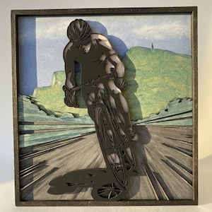 Puede incluir: Una obra de arte de madera en capas representa a un ciclista en movimiento sobre una carretera. El ciclista es de color gris oscuro, con casco y gafas de sol. El fondo presenta una colina verde y un cielo azul. La obra está enmarcada.