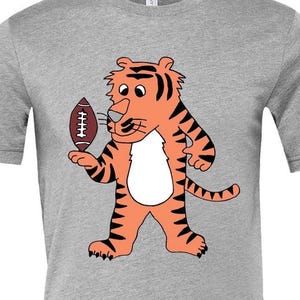 May include: A heather gray t-shirt featuring a cartoon tiger holding a football. The tiger is orange with black stripes and a white belly. The football is brown with white laces. The shirt is made of a soft material.