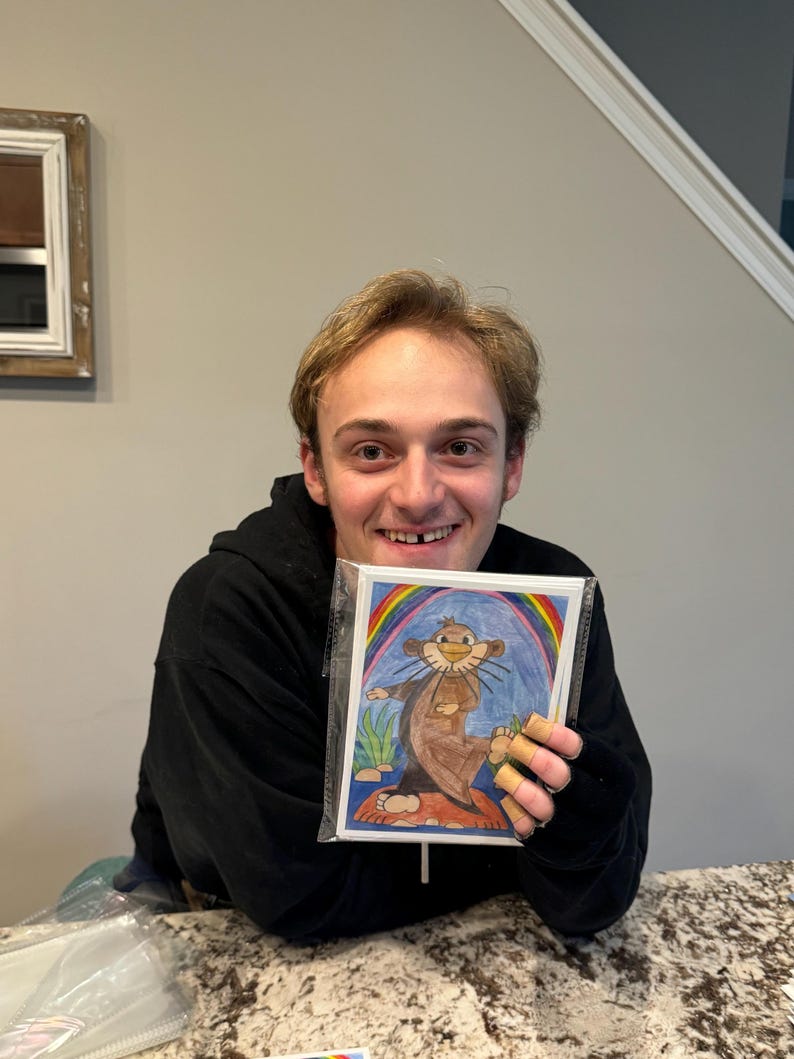 Puede incluir: Un joven sosteniendo un dibujo de una nutria de dibujos animados de pie sobre un hongo con un arco&iacute;ris detr&aacute;s.