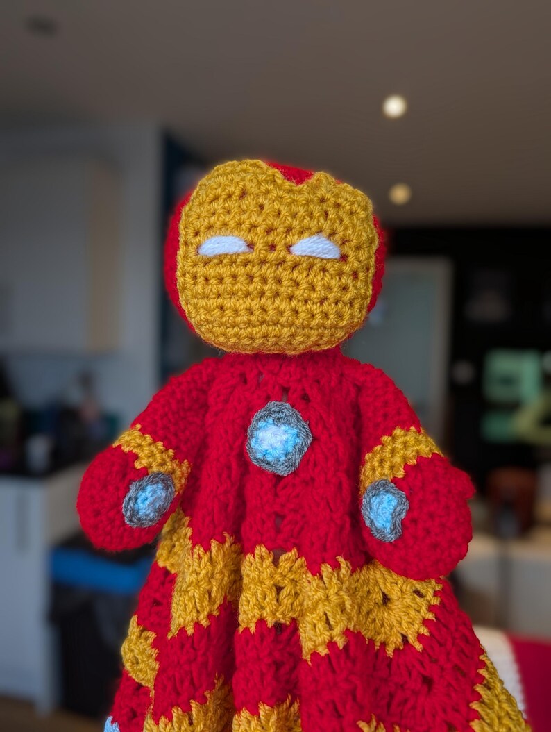 May include: A handmade crochet doll featuring a red and gold color scheme. The head has a gold faceplate with white eyes. The body includes a central light blue circular accent on the chest and matching light blue accents on the hands.