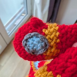 May include: A crocheted item featuring a vibrant red base with textured orange sections and a circular detail composed of a light blue center surrounded by a dark gray border, crafted with thick yarn in a dense, interlocking stitch pattern.