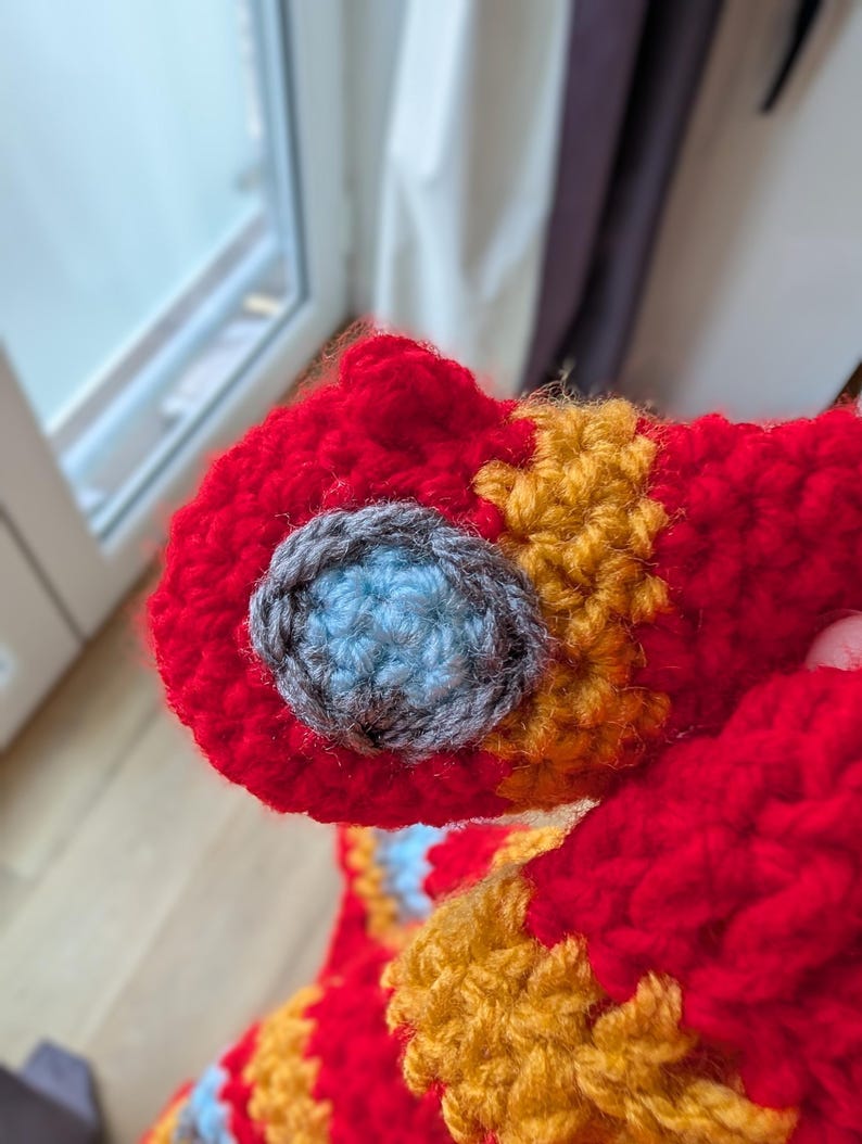 May include: A crocheted item featuring a vibrant red base with textured orange sections and a circular detail composed of a light blue center surrounded by a dark gray border, crafted with thick yarn in a dense, interlocking stitch pattern.