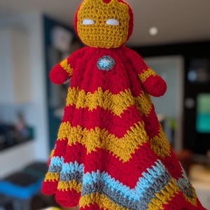 May include: A handmade crochet lovey featuring a stuffed head resembling a red and gold helmet with white eyes. The body is a soft, textured blanket with a chevron pattern in alternating stripes of red, gold, light blue, and gray yarn.